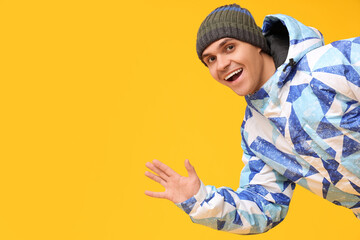 Young man in stylish puffer jacket and hat waving hand on yellow background