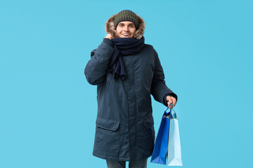 Young man in stylish puffer jacket and scarf with shopping bags on blue background