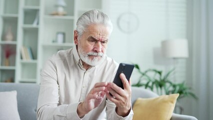 Frustrated elderly senior man having problems with software or service on smartphone sitting on sofa in living room at home. Worried disappointed mature male complains about bad internet connection
