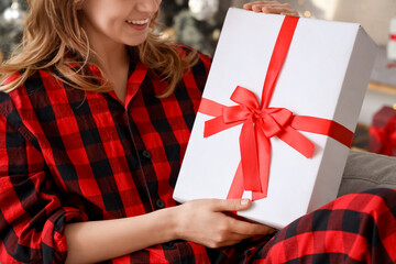 Happy young woman in pajamas with Christmas gift sitting at home, closeup