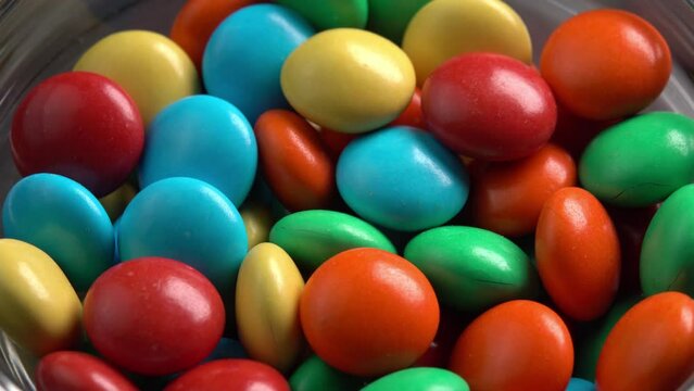 Sweet dragees in colored glaze are rotating in a glass bowl macro side light. Delicious sweets and candies close-up that children like. Chocolate Coating