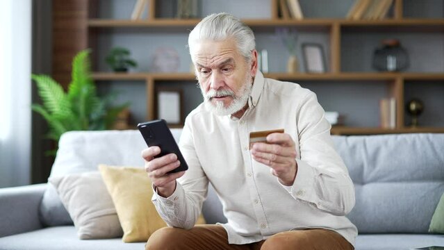 Frustrated elderly senior man discovered fraud while entering credit card number on smartphone sitting at home. Upset mature male spreads his hands and looks at camera. He became a victim of deception