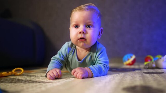 Little baby boy with fair hair and big eyes looks with interest around. Infant kid lies on the floor at home.