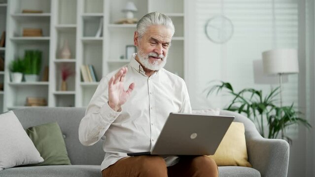 Handsome elderly senior gray haired man talking on a video call using a laptop computer while sitting on sofa in living room at home. Smiling positive adult mature male has online chat, remote meeting