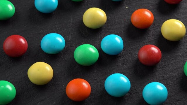 Sweet dragees in colored glaze rotating on black plate background macro top view low light. Delicious sweets and candies close-up that children like. Chocolate Coating