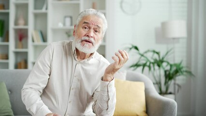 Webcam view. Elderly senior man having a video call with a doctor or psychologist sitting on sofa in room at home. Mature male telling about his problems, complaining about illness looking at camera