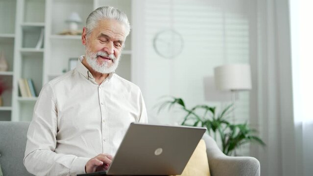 Elderly senior man working on laptop sitting on sofa in living room at home. Mature businessman texting messaging on computer browsing surfing web internet, online shopping, chatting, banking in app