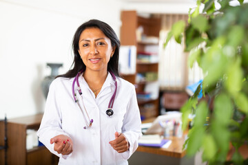 Positive hispanic female doctor assistant standing in medical office making welcoming gesture