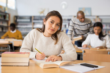 Portrait of positive young hispanic woman sitting at table in public library. Adult self-learning concept