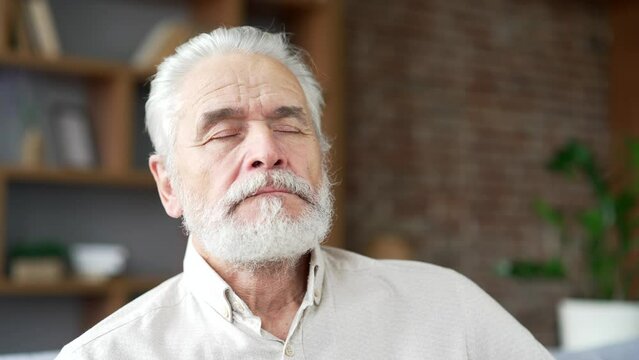 Elderly senior man with gray hair meditating eyes closed sitting on sofa in living room at home. Calm happy adult mature male pensioner relaxes at leisure, practices yoga breathing deeply. Close up