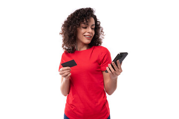 Naklejka premium 30 year old slim brunette woman dressed in a red basic t-shirt holds a smartphone and a credit card with a mockup