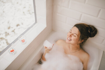 Young woman taking bath with foam near big window.