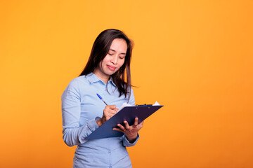 Serious asian woman writing business report on documents, analyzing questionnaire and answering question in studio. Confident model standing over yellow background looking at checklist - obrazy, fototapety, plakaty