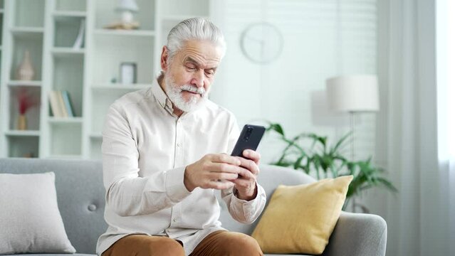 Elderly senior man with gray hair browsing smartphone sitting on sofa in living room at home. Adult mature pensioner reads, writes messages, chats, does online shopping, uses an application on phone