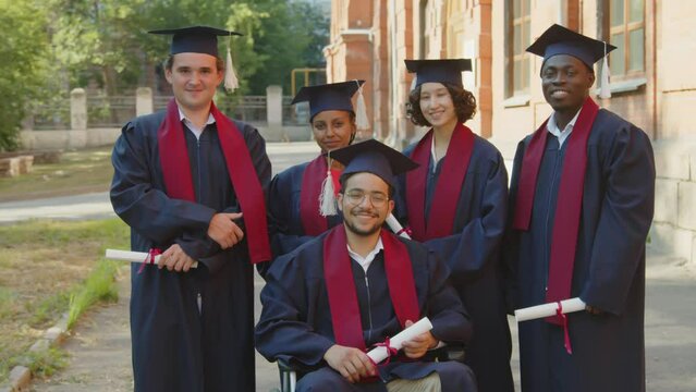 Portrait of diverse group of graduated students in gowns and hats standing next to their friend in wheelchair posing for photo