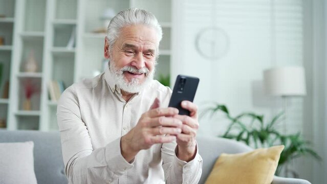Happy elderly senior man with gray hair received great news on smartphone while sitting on sofa in living room at home. Smiling adult mature male reads a pleasant message on phone, celebrates success