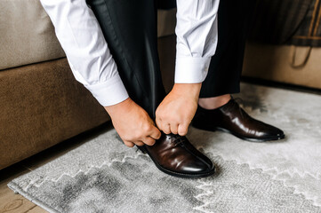 A man, the groom puts leather stylish shiny brown shoes on his feet in socks in the morning, sitting on the sofa and tying his shoelaces with his hands. Closeup photo of a businessman, business. - obrazy, fototapety, plakaty