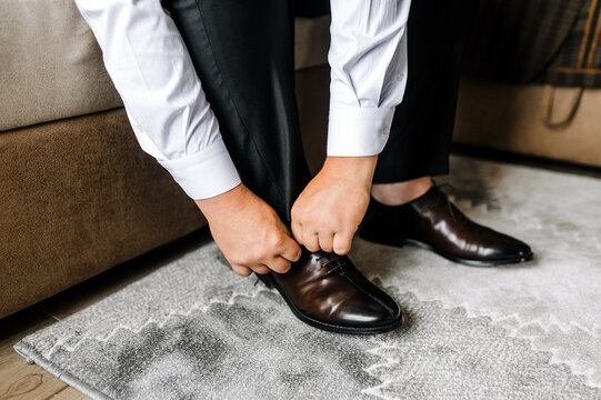 A man, the groom puts leather stylish shiny brown shoes on his feet in socks in the morning, sitting on the sofa and tying his shoelaces with his hands. Closeup photo of a businessman, business.