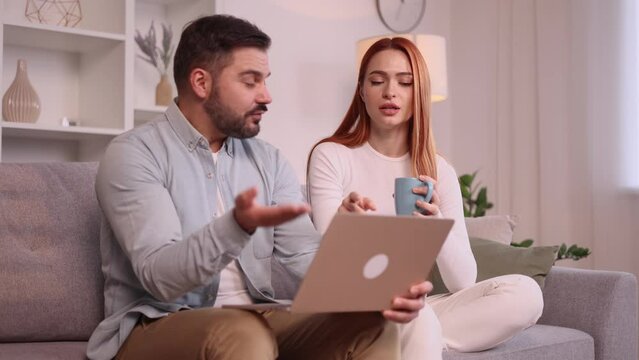 Young family couple looking at laptop computer screen choose products in online internet store make order online booking or plan trip emotionally discussed sitting on couch at home indoors 