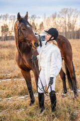Beautiful blond professional female jockey standing near horse in field in winter. Friendship with horse