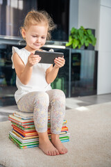 Cute Little girl sits on stack of children's fairy-tale books and watches cartoons on smartphone