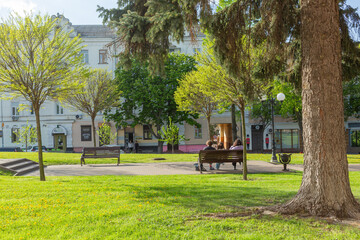 couple sitting on a bench in park with green trees and grass near red square in chernihiv