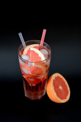 Grapefruit refreshing juice with ice, straws and pieces of citrus in a tall glass on a black background.