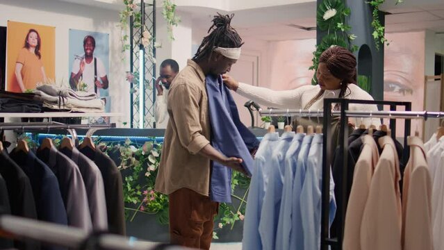 Couple in thrift shop looks at clothes, trying to find well fitting special occasion suits. Woman satisfied by cheap and high quality blazers in SH clothing store, giving ok hand sign approval gesture