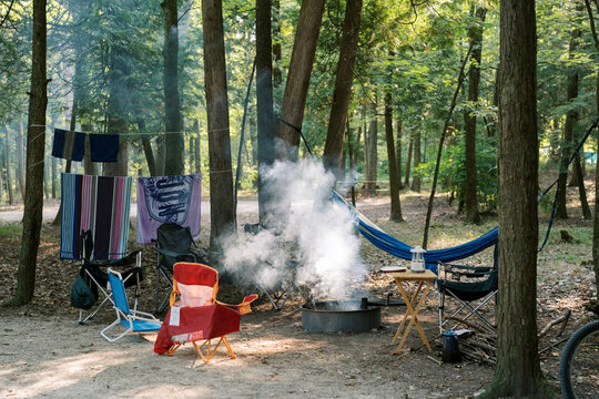 A horizontal image of a peaceful campsite with lawn chairs, campfire, and hammock in the forest. 