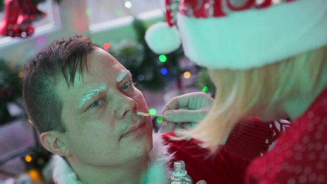 Make-up of young man in image of Santa Claus, close-up of girl applying glue to his face to add mustache and beard, preparing for Christmas holidays and New Year against backdrop of festive decoration
