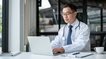 Professional senior Asian male doctor is working on his laptop computer in his office at the clinic.