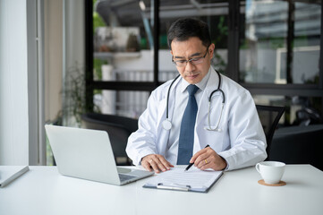 An experienced Asian senior male doctor is examining medical cases on report at his desk.