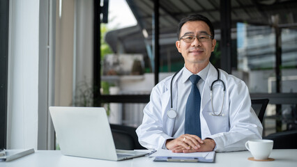 A successful and experienced senior Asian male doctor in a white gown is sitting at his desk.