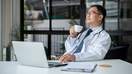 Fototapeta na wymiar Professional Asian senior male doctor is sipping coffee while working in his office at the hospital.