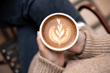 A woman in a cosy knitted sweater holding a cup of hot cappuccino, enjoying morning coffee at cafe.