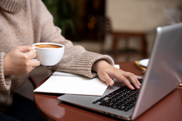 A woman is working remotely at a cafe, sipping hot coffee while working on her laptop.