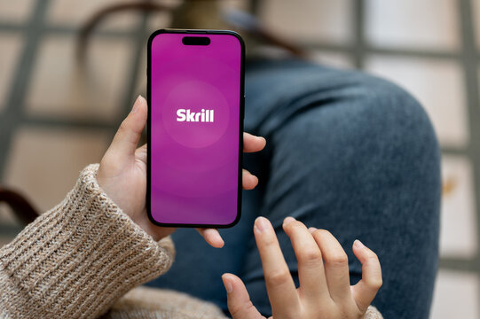 A woman using Skrill app on her IPhone14 while sitting outdoors. Skrill logo on IPhone14 screen.