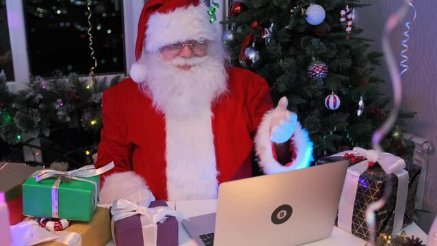 Close up man in Santa Claus costume sitting at laptop, using keyboard, reading emails with children wishes, next to gifts boxes, candles, Christmas tree, branches, garlands. Xmas time.