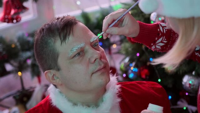 Male actor is preparing to transform into Santa, makeup artist applies glue and paint to his face before Xmas party, against backdrop of festive tree and glowing garlands. New Year holiday prepare.
