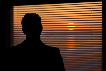 A man stands in a room and looks out the window at the setting sun.