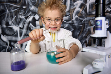 Cute blond boy participates in scientific study and conducts chemical scientific experiment by analyzing and mixing liquid in test tube. Boy mixes substances different colors in cones different shapes