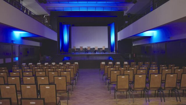 Empty conference room with empty chairs on stage - sow motion dolly shot. Seminar in the old building with blue lights. Stage with no people in audience ready for meeting. Events during covid period.