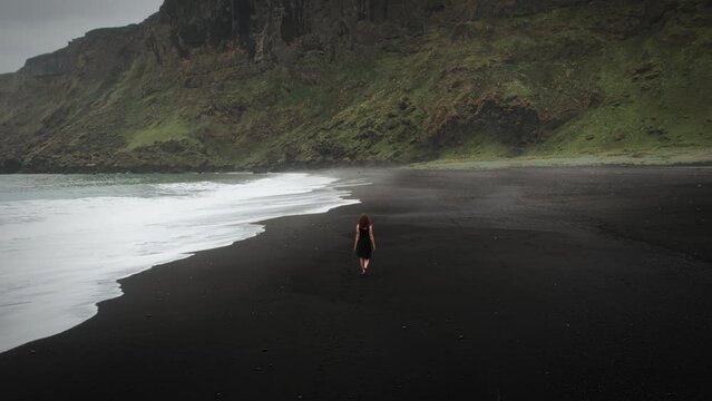 Young beautiful redhead woman walking along a black sand beach barefoot in a black dress. Cloudy day Vik, Iceland with epic dark green mountains and ocean waves in distance. Aerial drone 4K