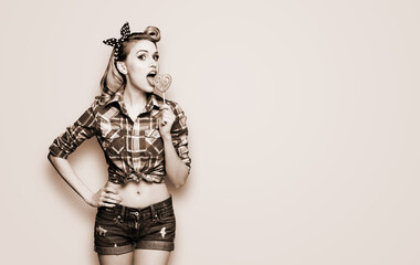 Portrait of beautiful woman licking heart shape lollipop dressed in pinup plaid shirt. Pin up girl...