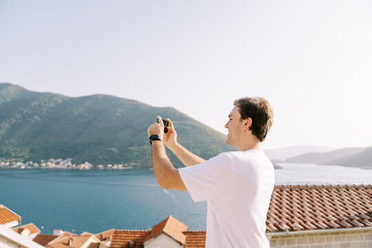 Young man takes a photo with a smartphone of a view of the mountains and the sea over the red roofs of houses