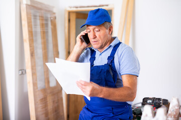 Worried middle-aged male repairer in a blue overalls calling on a mobile phone while holding a work plan while renovating a house