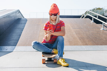 Beautiful young hipster woman using smart phone On Skateboard at summer day. Active lifestyle, modern life, subculture concept