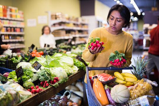 Portrait of pensive latin american woman buying fresh organic vegetables in supermarket, choosing red radish