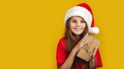 Happy little girl in Christmas hat holding present box isolated on yellow background