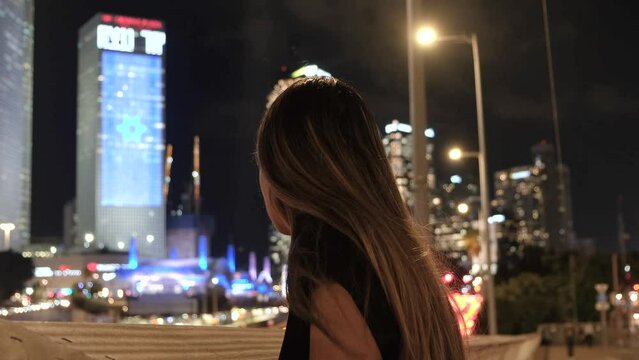 Night shot of huge Israel flag hanging on Azrieli tower with a young concerned jewish woman looking at it from a bridge. Big city lights shot of a worried lady with a focus on Israel flag background.